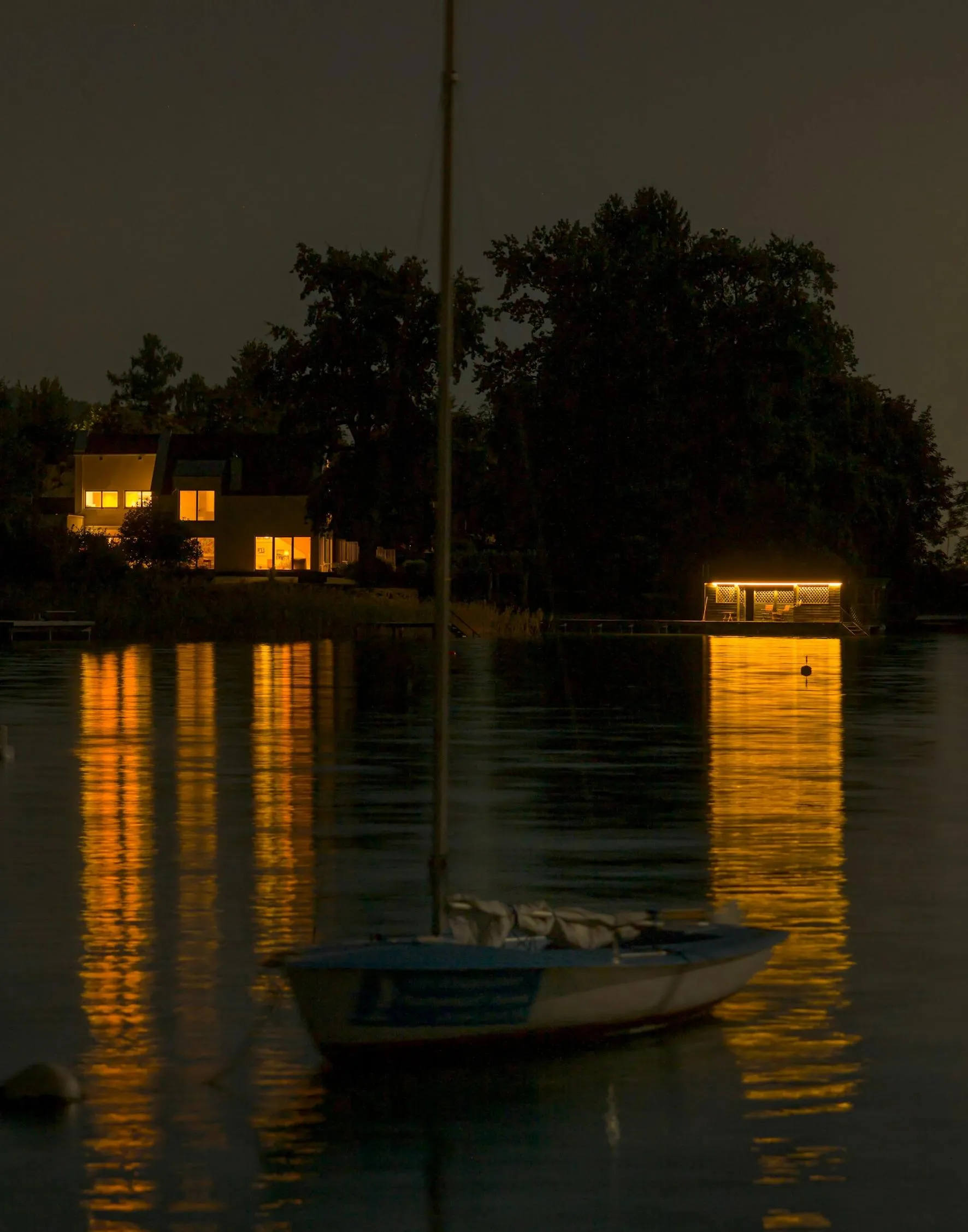 A still boat in the middle of a lake; sunset-like house lights in the backdrop.