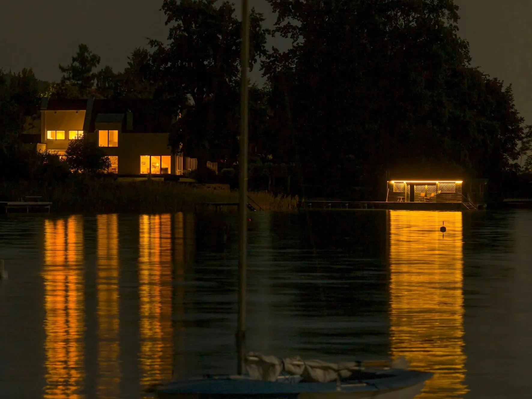 A still boat in the middle of a lake; sunset-like house lights in the backdrop.